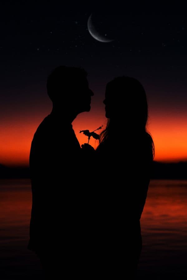 Couple silhouetted against a red sunset under a crescent moon